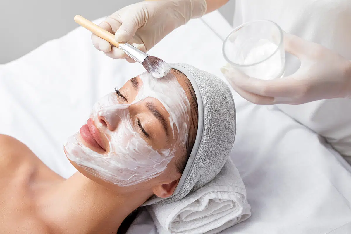 beautician applying white clay mask to face of beautiful young woman