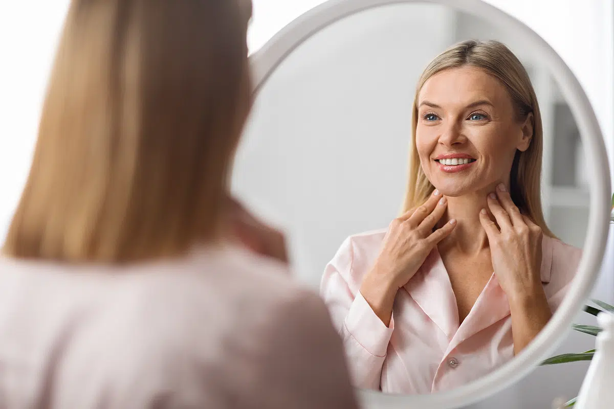 beautiful mature woman looking at mirror and touching soft skin on neck