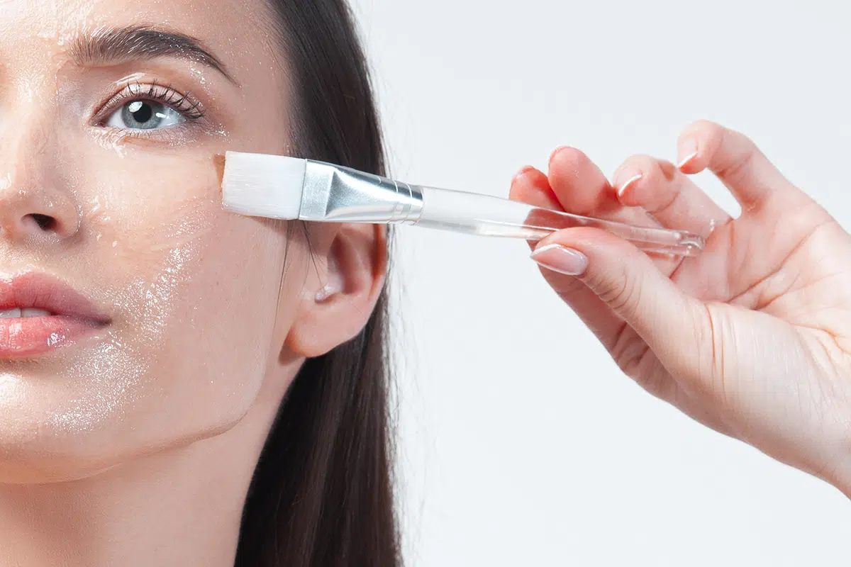young brunette woman applying moisturizing gel mask with brush on her face